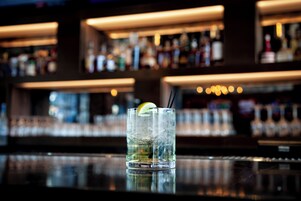 Um bar com um copo de bebida gelada, uma fatia de limão e canudo, e prateleiras com garrafas ao fundo.