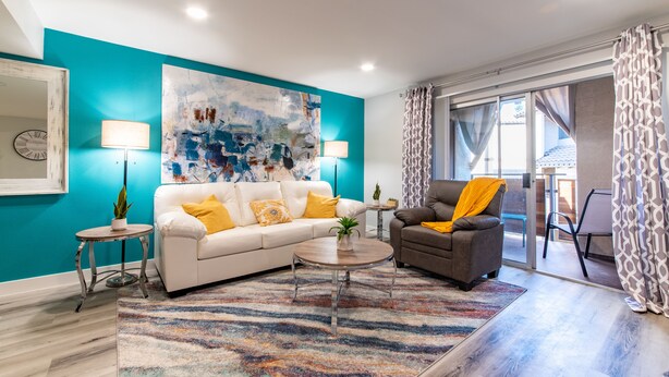 Una sala moderna con un sofá blanco, un sillón gris y una alfombra colorida. Hay un cuadro abstracto grande en la pared y un reloj a la izquierda.