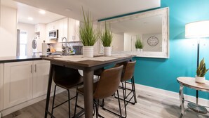 Una cocina moderna con armarios blancos, una mesa de comedor de madera y sillas marrones. Hay plantas en macetas sobre la encimera y un reloj en la pared.