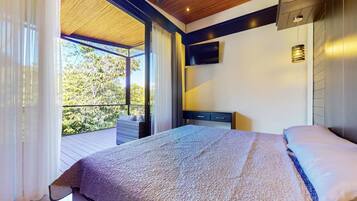 A bedroom with a large bed, a nightstand, a TV, and a view of greenery outside.