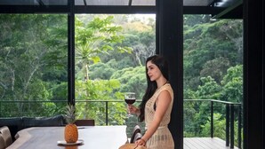 A woman standing by a table with a pineapple and a glass of red wine, overlooking a lush forest view from a modern interior with large windows and a balcony. - Odyssey Villa - incredible mountain views, 3 bedrooms, w pool! (Peréz Zeledon)