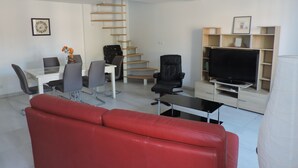 Una sala moderna con un sofá rojo, una mesa de comedor con sillas, una silla de oficina negra, un televisor sobre un mueble bajo y una escalera de caracol.