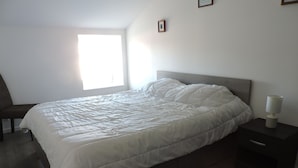 Una cama bien hecha con edredón blanco, una lámpara de noche y una silla en una habitación sencilla con ventana.