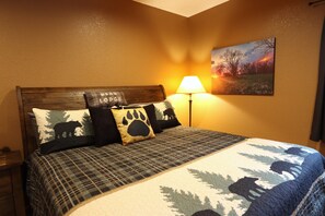 Cama com manta xadrez e três travesseiros, um com desenho de urso, em quarto com pintura de pôr do sol e abajur na mesinha de cabeceira.