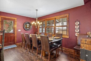 Uma sala de jantar com mesa e cadeiras de madeira, um candelabro e grandes janelas com vista para árvores.