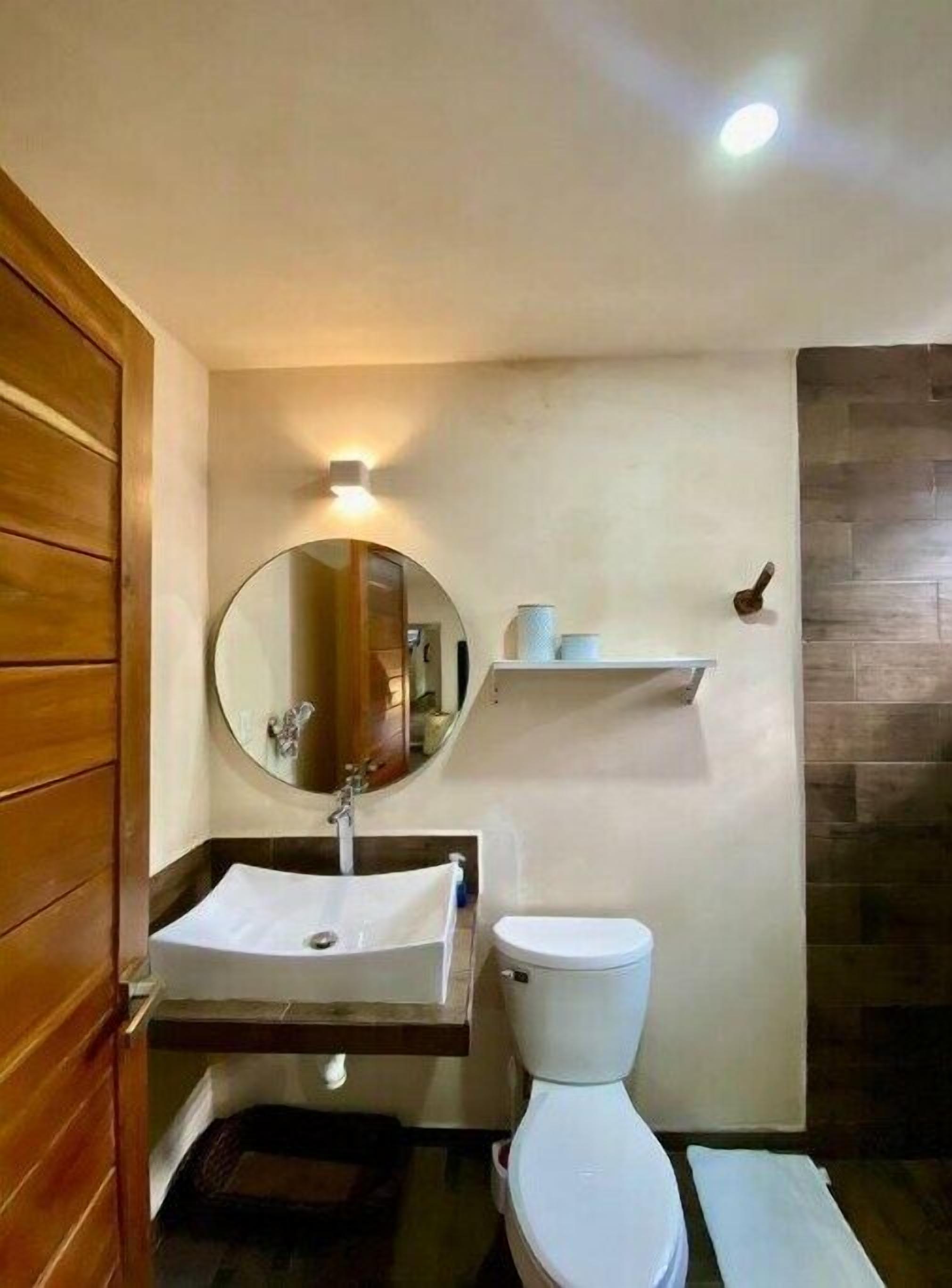 A bathroom with a wooden door, a round mirror, a white sink, a white toilet, and a shelf with toiletries.