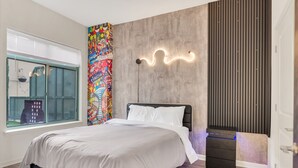 Une chambre moderne avec un lit, une fenêtre, une fresque murale colorée et un luminaire mural original.