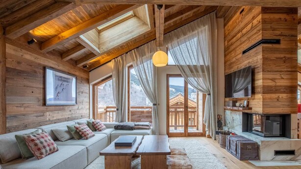 L’intérieur chaleureux d’un chalet en bois comprend un canapé, des tables en bois, une cheminée et de grandes fenêtres qui offrent une vue sur les montagnes.