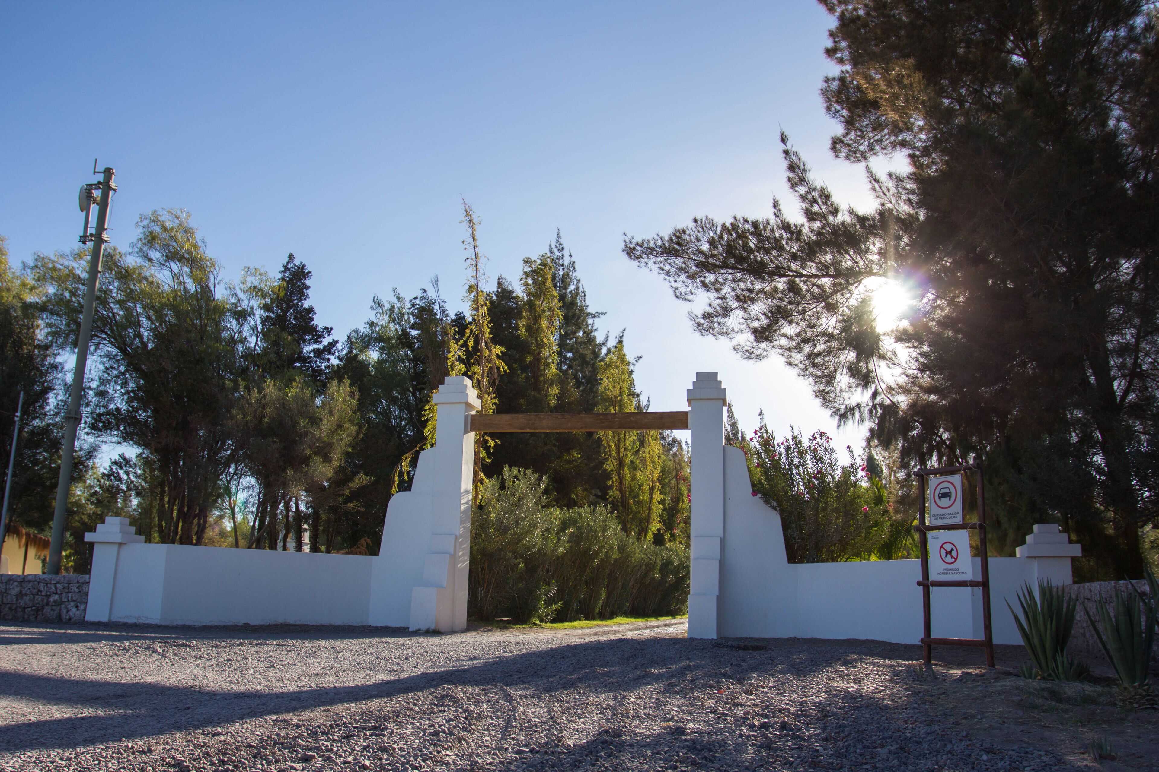 A gravel driveway leading to a white gate with a wooden archway, surrounded by trees and a clear sky.