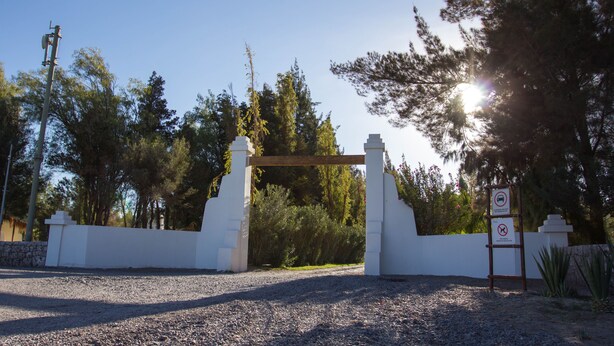 A gravel driveway leading to a white gate with a wooden archway, surrounded by trees and a clear sky. - Cabañas La Huayca - Caja Los Andes (Pozo Almonte)