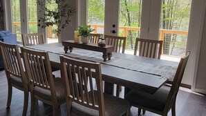 A dining area with a large wooden table and chairs, surrounded by large windows offering a view of trees and a wooden deck outside. - Clover House at Alpine Lake with pool access. (Terra Alta)