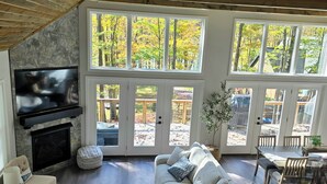 A living room with a stone fireplace, a large TV, a sofa, and a dining area with a view of trees. - Clover House at Alpine Lake with pool access. (Terra Alta)