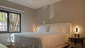 Ein modernes Schlafzimmer mit einem großen Bett, einem Nachttisch und einem Fenster mit Blick auf Bäume.