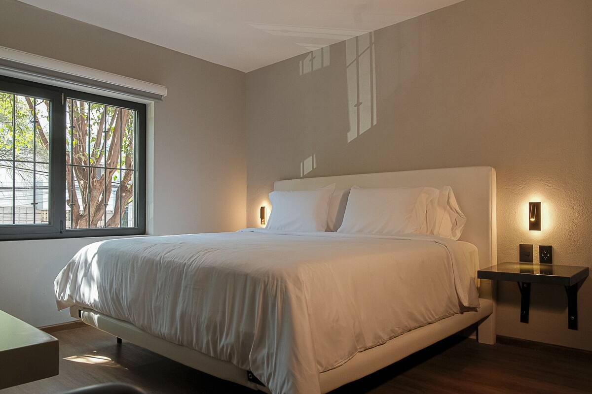 A modern bedroom with a large bed, a bedside table, and a window with a view of trees.