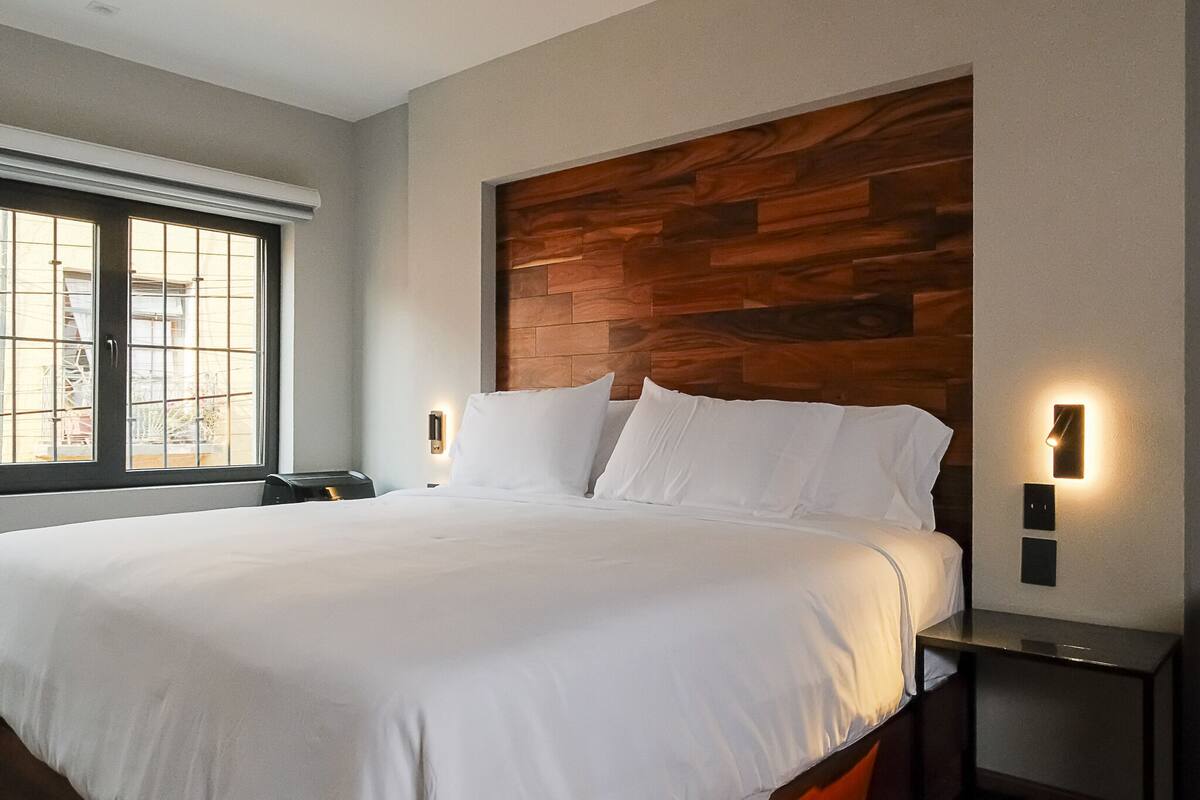 A modern hotel room with a large bed, wooden headboard, two bedside tables with wall-mounted lights, and a window with blinds.