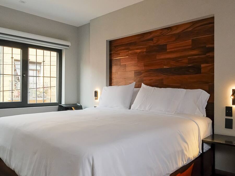 Habitación de hotel moderna con una cama grande, cabecera de madera, dos mesitas de noche con lámparas fijadas a la pared y una ventana con persianas.