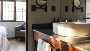 Ein Hotelzimmer mit einem hölzernen Schminktisch, einem runden Spiegel, einem weißen Waschbecken und einem grauen Sessel.