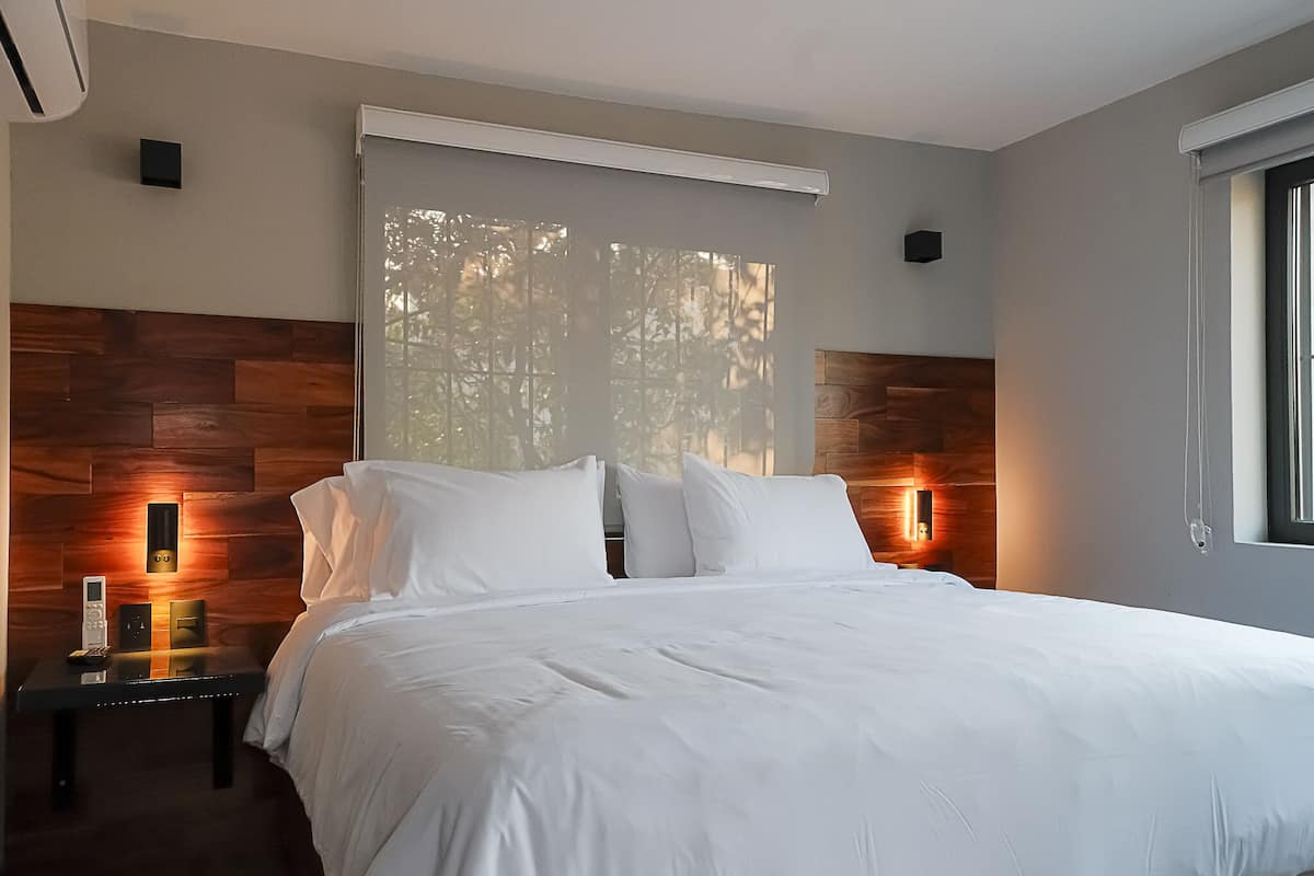 A modern bedroom with a large bed, wooden headboard, bedside lamps, and a window with a view of trees.