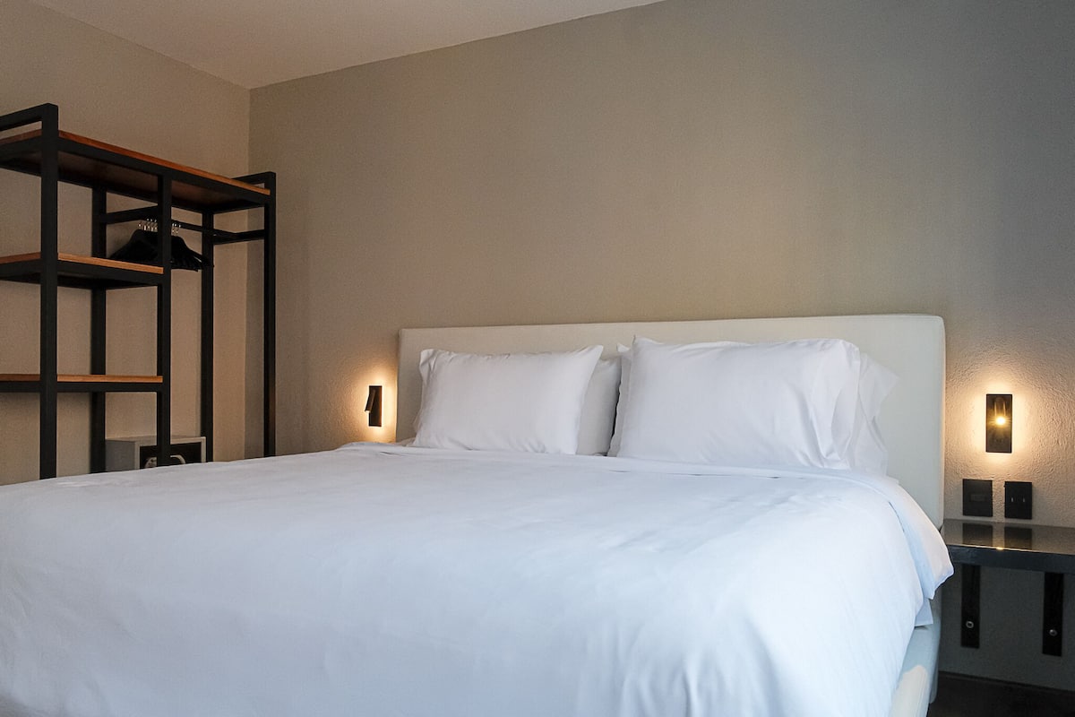A neatly made bed with white linens, a wooden shelf unit, and two wall-mounted light fixtures.