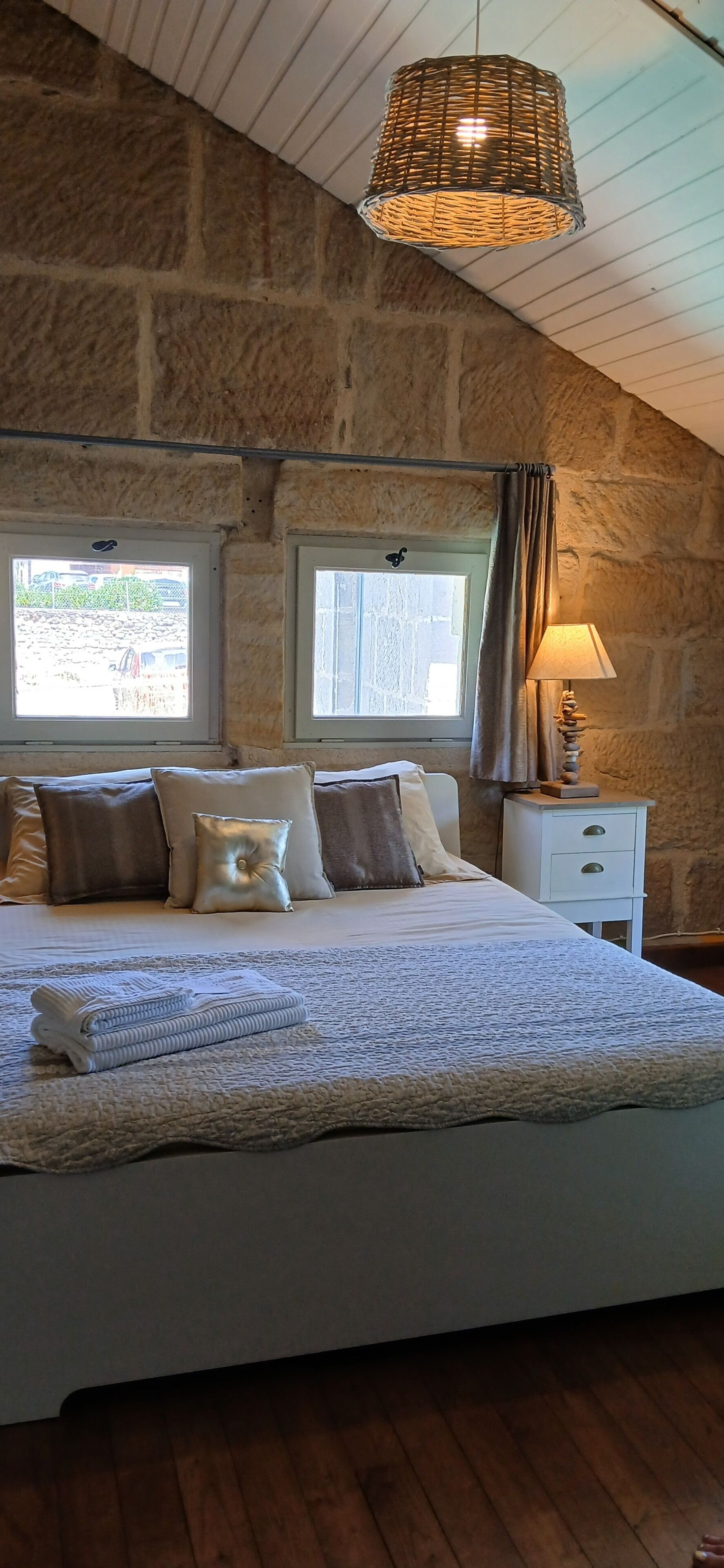 Une chambre avec un mur en pierre, un lit avec des oreillers, une lampe de chevet et des fenêtres.