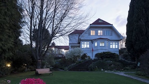 Een groot, twee verdiepingen tellend huis met een rood dak, omgeven door een verzorgde tuin en een bankje.