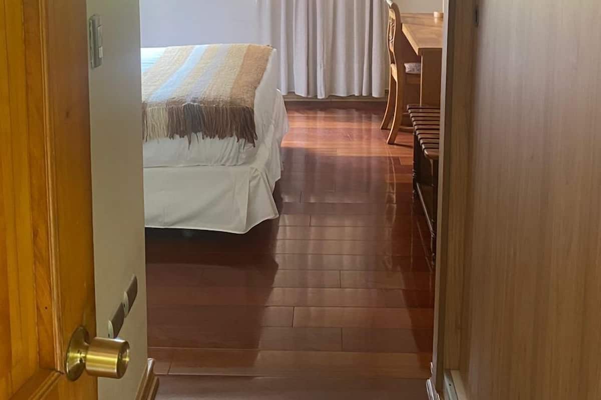 Une chambre d’hôtel avec un lit, un bureau et une chaise. Il y a une fenêtre avec des rideaux, et une porte est visible sur la gauche.