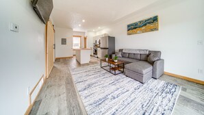 En moderne stue med en grå sofa, et sofabord og en blå og hvid måtte. I baggrunden ses et køkken med hvide skabe og et gråt køleskab.