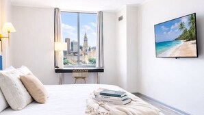 Une chambre d’hôtel avec un grand lit, une vue sur la ville par la fenêtre, un téléviseur à écran plat fixé au mur et une petite table avec une chaise.