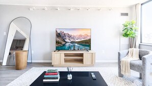 Un salon moderne avec un téléviseur à écran plat posé sur un meuble en bois, une table basse avec des livres et une télécommande, un fauteuil gris et une grande fenêtre agrémentée d’une plante.