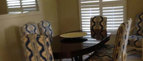 A dining area with a round table surrounded by six patterned chairs, a chandelier above, and large windows with blinds.
