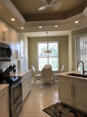 A modern kitchen with white cabinets, a built-in oven, a microwave, and a dining area with wicker chairs and a glass table. - Adorable Floridian Rental (Naples)
