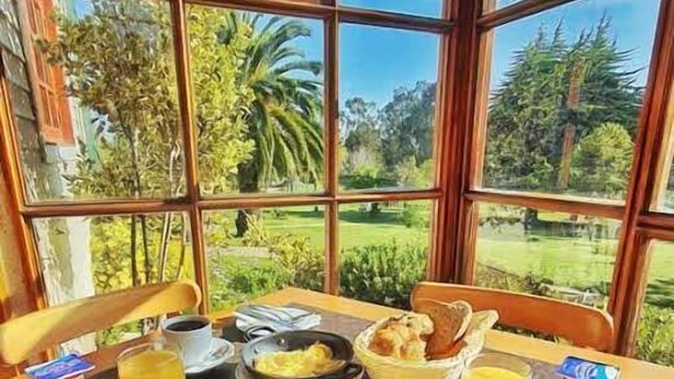 A breakfast table with a variety of food items and utensils, set against a backdrop of large windows overlooking a garden. - Hotel y Cabañas Huallilemu Sur- Caja Los Andes (El Quisco)