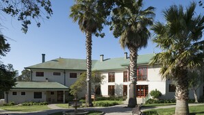 A two-story building with a green roof, surrounded by palm trees and a paved walkway. - Hotel y Cabañas Huallilemu Sur- Caja Los Andes (El Quisco)