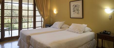 A hotel room with two beds, a large window with curtains, a framed artwork on the wall, and a bedside table with a phone.