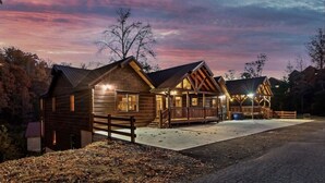 A wooden cabin with a porch and a covered deck, surrounded by trees and a gravel driveway.