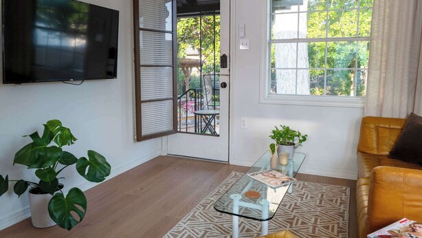 A living room with a glass coffee table, a sofa, a TV mounted on the wall, and a door leading to an outdoor area.