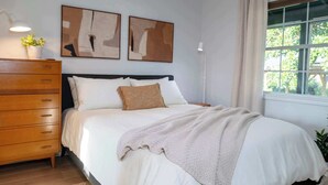 Ein Schlafzimmer mit einem Bett, einer Holzkommode, zwei abstrakten Gemälden, einem Fenster mit Vorhängen und einer Topfpflanze.