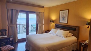 Quarto de hotel com uma cama grande, uma escrivaninha, uma cadeira e uma varanda com cortinas.