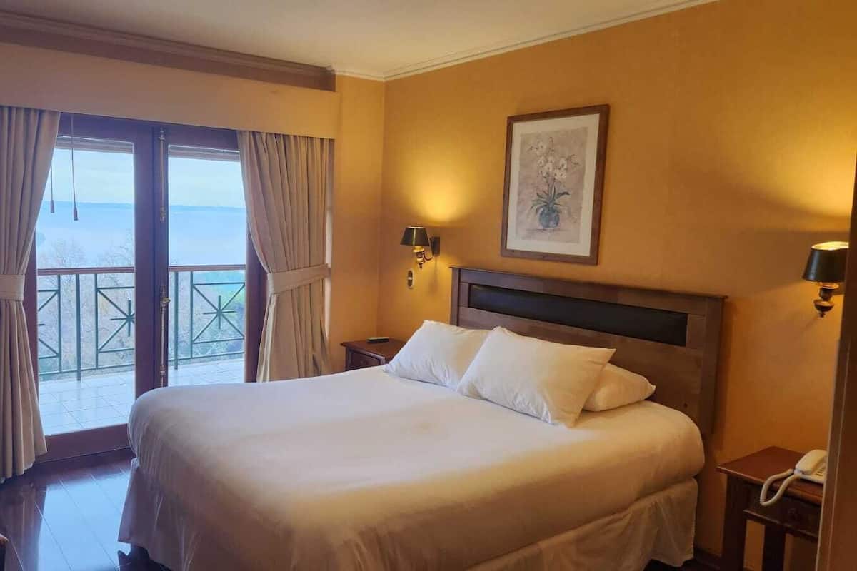 A hotel room with a large bed, a desk, a chair, and a balcony with curtains.