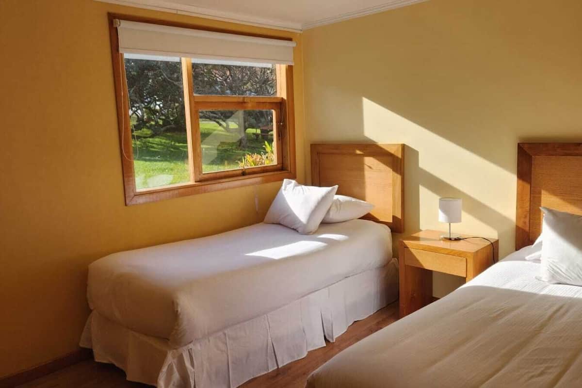 A bedroom with two single beds, a window with a view of greenery, and a bedside table with a lamp.