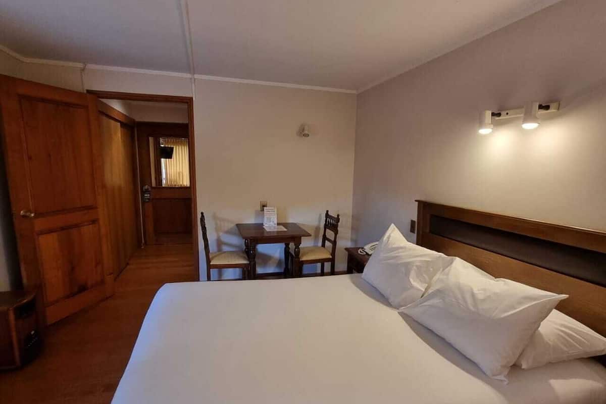 A hotel room with a large bed, a wooden headboard, a small table with chairs, and a wooden wardrobe.