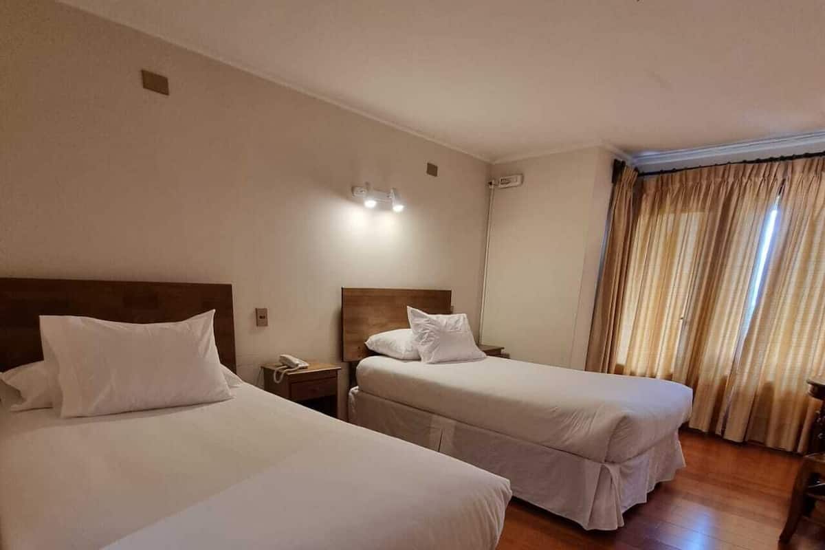 A hotel room with two beds, wooden flooring, a nightstand with a phone, and a window with curtains.