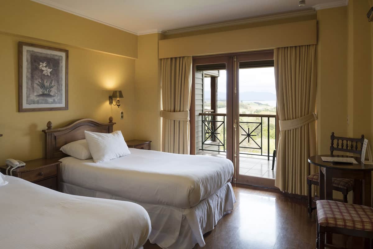 A hotel room with two beds, a desk, a chair, and a balcony with curtains.