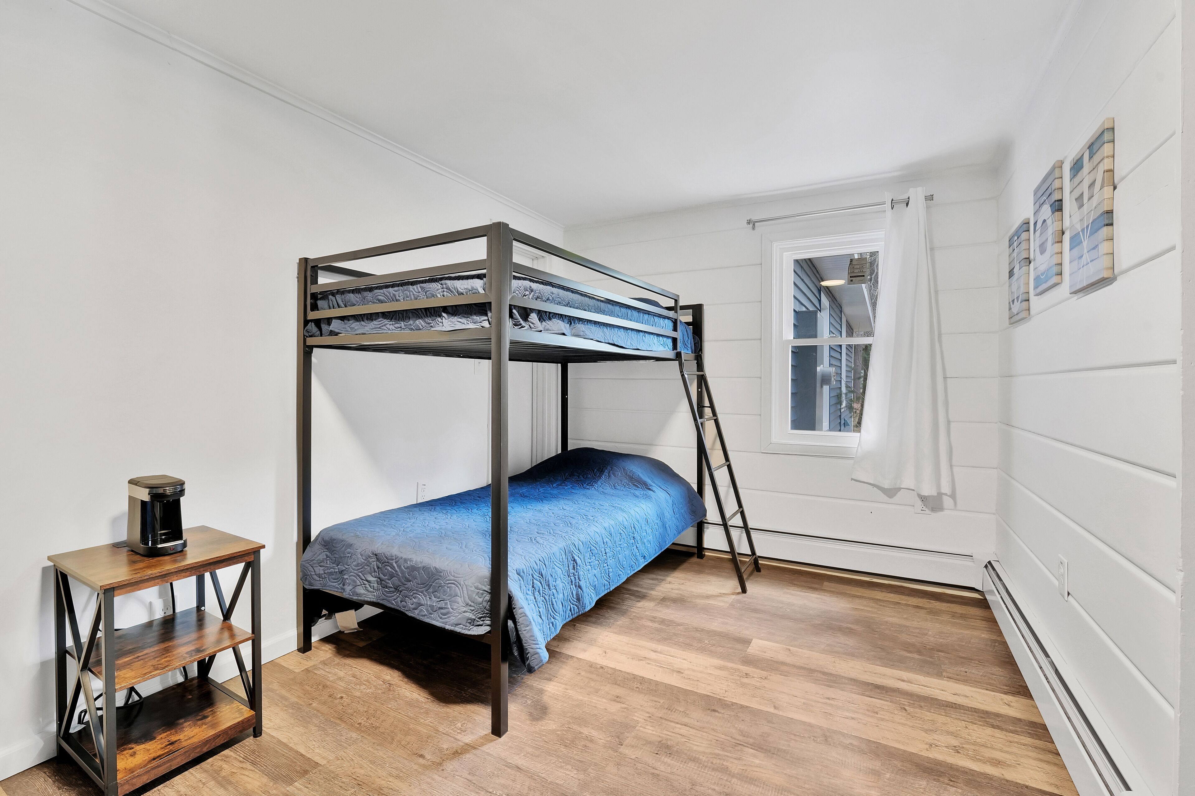 Un lit superposé avec un matelas bleu, une table de chevet en bois sur laquelle se trouve une machine à café, et une fenêtre avec des rideaux.