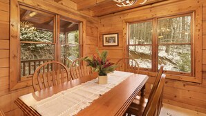 Interior de una cabaña de madera con una mesa de comedor, sillas, una araña y ventanas amplias que dan a un bosque.