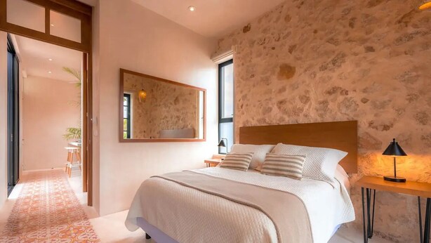 A bedroom with a large bed, a wooden headboard, a stone wall, and a small bedside table with a lamp. - Kinal 05 - One Bedroom Apartment, Sleeps 2 (Mérida)