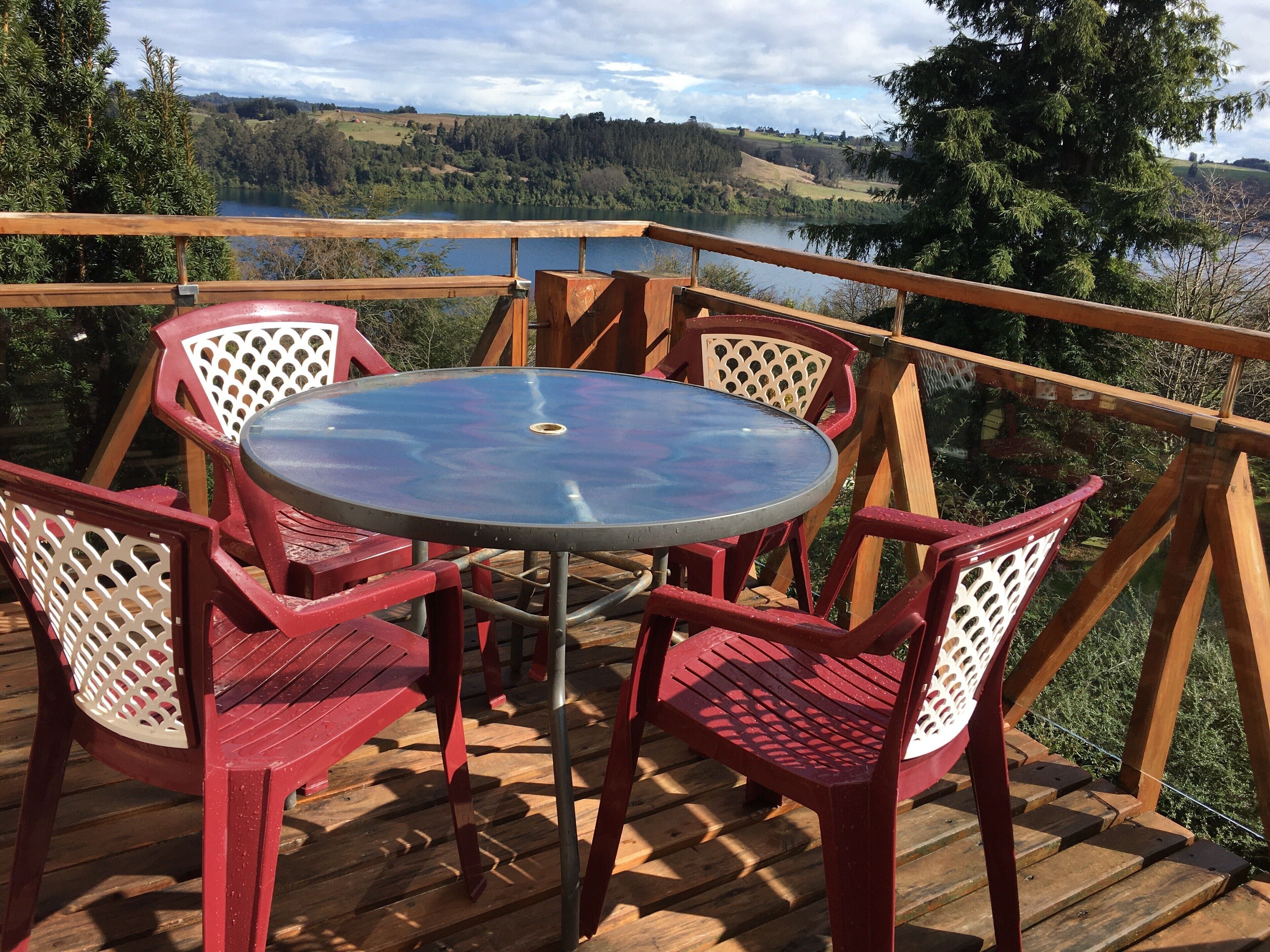 Una terraza de madera con una mesa de vidrio y sillas rojas, con vistas a un lago y colinas boscosas.