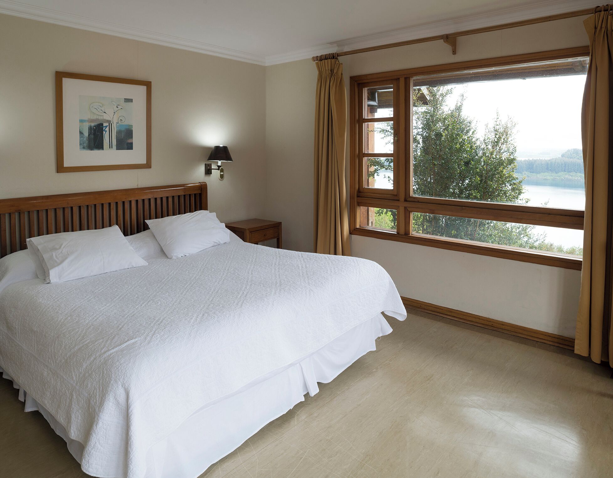 A neatly made bed with white linens in a room with a large window offering a view of trees and a lake.