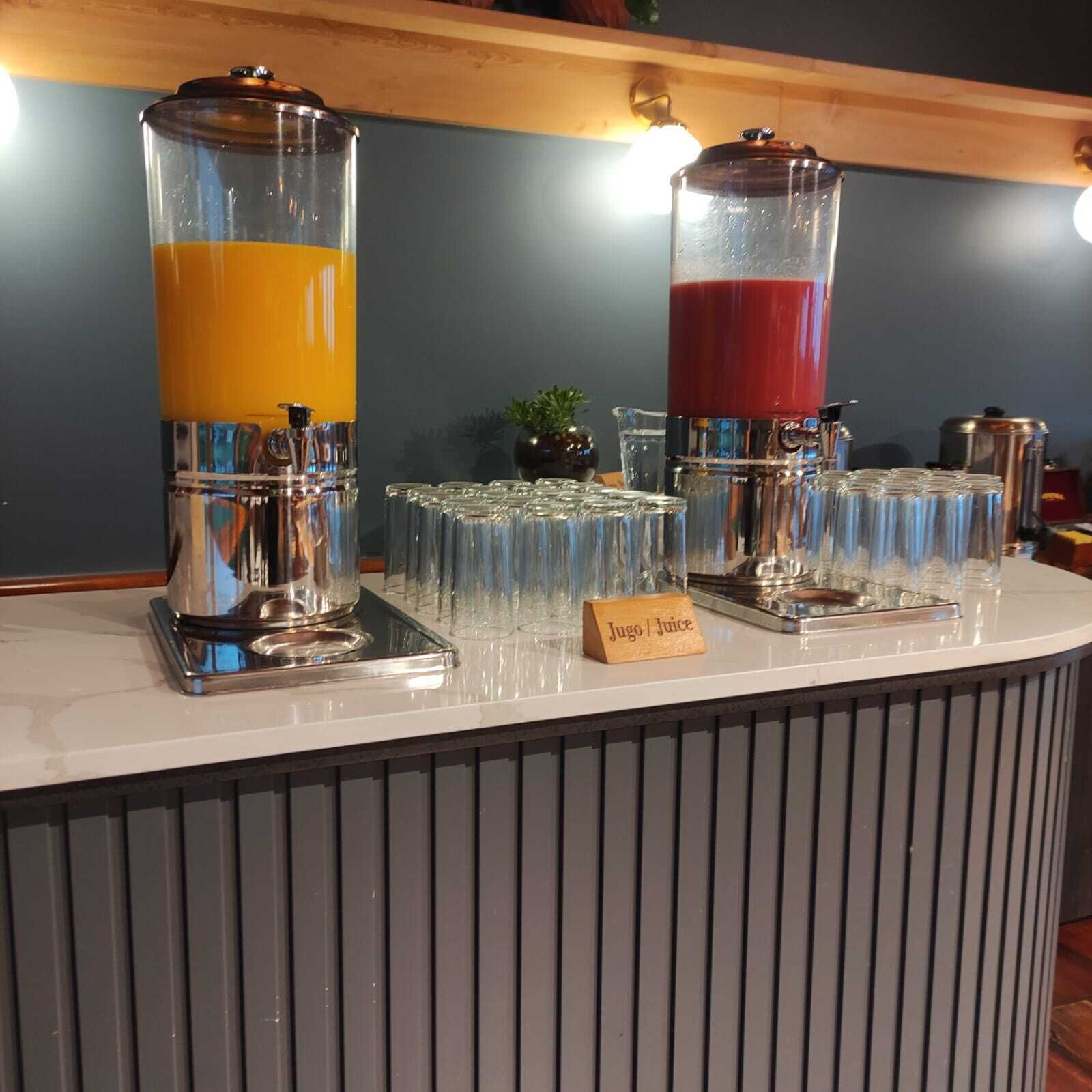 Un bar de sucs amb dos dispensadors, un per suc d'aranja i un altre per suc vermell, i un cartell que diu 'Jugo / Juice'.