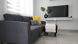 Ein modernes Wohnzimmer mit einem grauen Sofa, einem gelben Kissen, einem schwarz-weiß gestreiften Kissen, einer weißen Decke und einem kleinen schwarzen Hocker. An der Wand hängt ein großer Flachbildfernseher über einem weißen Regal mit einer Pflanze und Dekoration.